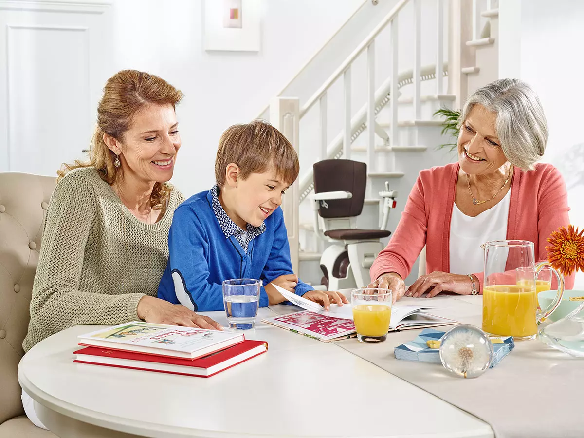 Sanitätskaufhaus Kellner - Relaunch 2025 - Kaufhaus - Treppenlifte - Familie sitzt am Esstisch im Wohnzimmer mit Treppenlift & Treppe im Hintergrund
