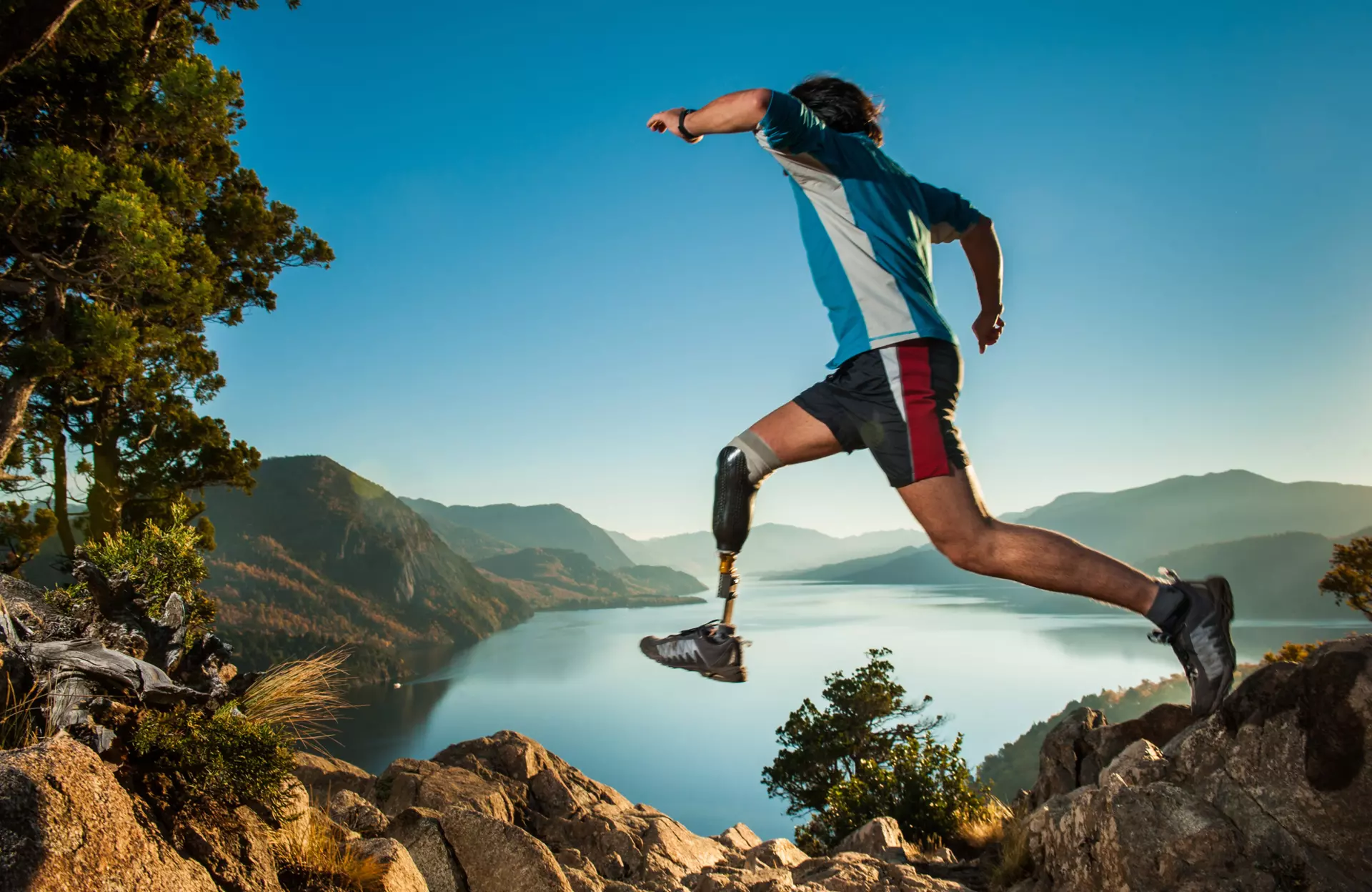 Sanitätskaufhaus Kellner - Relaunch 2025 - Stockmaterial - Sportler springt abenteuerlich mit Beinprothese von einem Stein zum anderen vor Bergen und einem großen See im Hintergrund